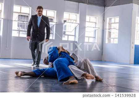Diverse competitors in blue white gis grappling on mats in martial hall with referee observing Diverse competitors in blue white gis grappling on mats in martial hall with referee observing 141539928