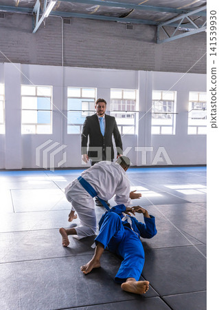 Diverse male martial artists in gis grappling in dojo while referee in suit watching Diverse male martial artists in gis grappling in dojo while referee in suit watching 141539930