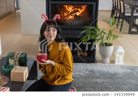 Hispanic woman wearing antler headband holding mug beside fireplace with wrapped gifts Hispanic woman wearing antler headband holding mug beside fireplace with wrapped gifts 141539949