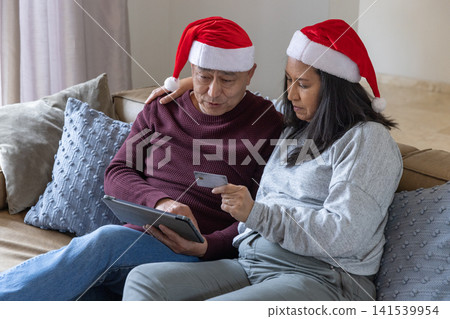 Diverse senior couple in santa hats sitting on sofa at home browsing tablet with credit card Diverse senior couple in santa hats sitting on sofa at home browsing tablet with credit card 141539954