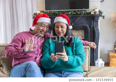 Diverse couple in santa hats holding smartphone and waving on sofa by fireplace mantel Diverse couple in santa hats holding smartphone and waving on sofa by fireplace mantel 141539955