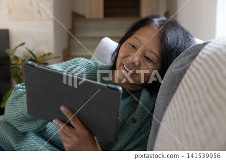 Senior asian woman reclining on cushioned sofa against pillow in living room holding tablet Senior asian woman reclining on cushioned sofa against pillow in living room holding tablet 141539956