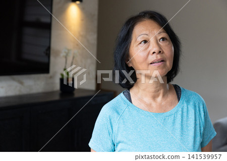 Senior asian woman gazing upward in living room in shirt beside orchid, copy space 141539957