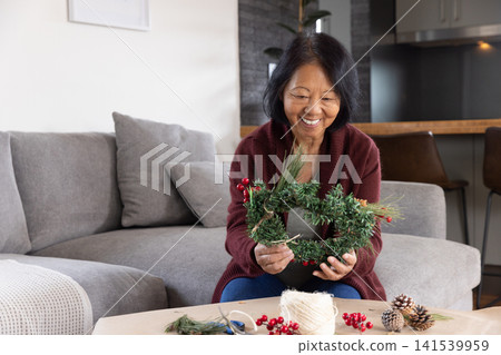 Senior Asian woman sitting in living room crafting wreath with pine cones, red berries, copy space Senior Asian woman sitting in living room crafting wreath with pine cones, red berries, copy space 141539959