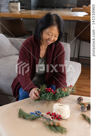 Senior asian woman assembling holiday wreath on coffee table at home with pine sprigs, berries Senior asian woman assembling holiday wreath on coffee table at home with pine sprigs, berries 141539960
