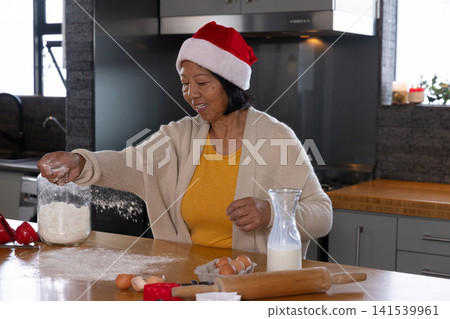 Asian senior woman in Santa hat sprinkling flour on countertop near eggs in kitchen, copy space Asian senior woman in Santa hat sprinkling flour on countertop near eggs in kitchen, copy space 141539961