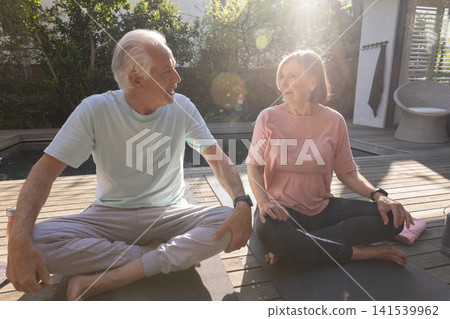 Senior couple practicing seated yoga on yoga mats on wooden deck next to pool with smartwatches 141539962