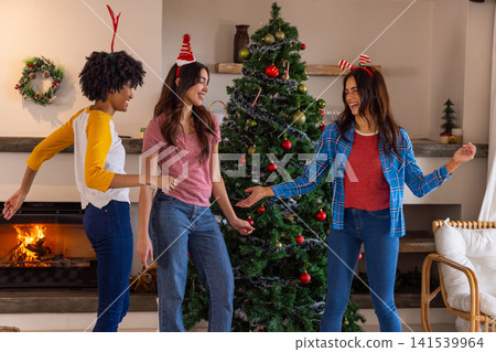Diverse female friends gathering around christmas tree with ornaments by lit fireplace 141539964
