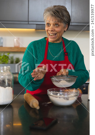 African American senior woman wearing red apron sifting flour with metal sieve on kitchen counter African American senior woman wearing red apron sifting flour with metal sieve on kitchen counter 141539977