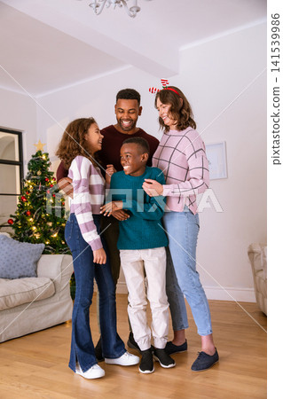 Diverse family hugging and smiling in living room near decorated christmas tree, chandelier Diverse family hugging and smiling in living room near decorated christmas tree, chandelier 141539986