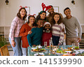 Diverse family standing with arms around one another at wooden table with salad plates, headwear 141539990