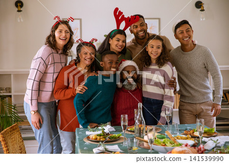 Diverse family standing with arms around one another at wooden table with salad plates, headwear Diverse family standing with arms around one another at wooden table with salad plates, headwear 141539990
