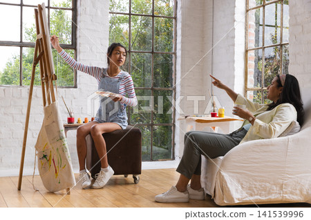 Asian teenage girl painting on canvas at easel in studio as African American instructor pointing Asian teenage girl painting on canvas at easel in studio as African American instructor pointing 141539996