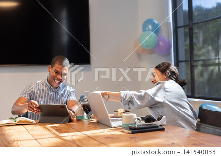 Diverse male coworkers collaborating on laptop at conference table with muffins, pastel balloons Diverse male coworkers collaborating on laptop at conference table with muffins, pastel balloons 141540033