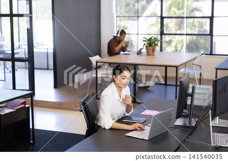 Diverse coworkers working in open office typing on laptop with coffee mug wearing headphones Diverse coworkers working in open office typing on laptop with coffee mug wearing headphones 141540035