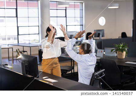 Diverse coworkers in business attire high-fiving beside desk in office with monitors Diverse coworkers in business attire high-fiving beside desk in office with monitors 141540037