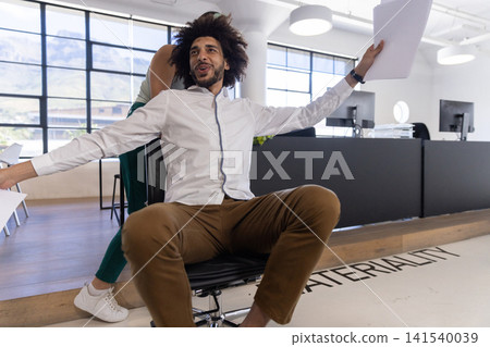 Diverse coworkers pushing rolling office chair and holding paper document in office interior Diverse coworkers pushing rolling office chair and holding paper document in office interior 141540039