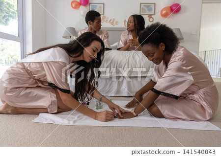 Diverse female friends kneeling in bedroom painting nails while sipping champagne by bridal sign Diverse female friends kneeling in bedroom painting nails while sipping champagne by bridal sign 141540043
