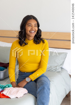 Woman sitting on bed at home packing folded clothes into open carry-on suitcase Woman sitting on bed at home packing folded clothes into open carry-on suitcase 141540046