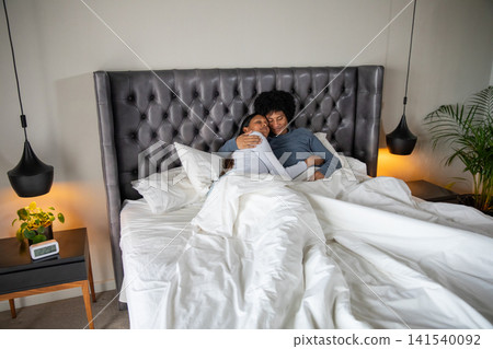 Diverse couple lying under white duvet on bed with tufted headboard, pendant lamps Diverse couple lying under white duvet on bed with tufted headboard, pendant lamps 141540092