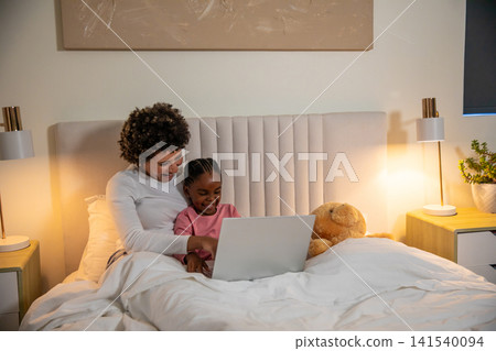 Diverse mother with daughter smiling while using laptop with teddy bear on bed with headboard Diverse mother with daughter smiling while using laptop with teddy bear on bed with headboard 141540094