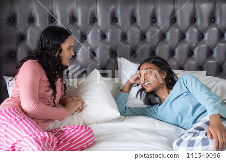 African american female friends relaxing and chatting on bed with headboard and pillows African american female friends relaxing and chatting on bed with headboard and pillows 141540096