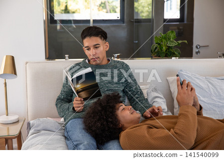 Diverse male couple reading sunset book and scrolling smartphone on bed in bedroom with gold lamp Diverse male couple reading sunset book and scrolling smartphone on bed in bedroom with gold lamp 141540099