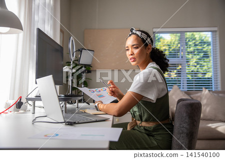 Woman holding printed chart, pen while studying data on laptop at home office corner 141540100