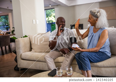 Diverse senior couple taking blood pressure reading on sofa and high-fiving with monitor Diverse senior couple taking blood pressure reading on sofa and high-fiving with monitor 141540105
