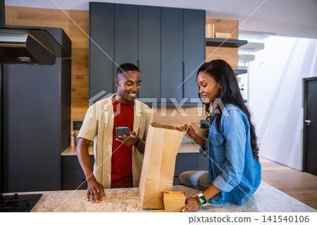 African American couple unpacking groceries from bag at kitchen island while checking smartphone African American couple unpacking groceries from bag at kitchen island while checking smartphone 141540106