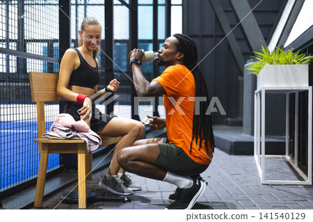 Diverse training partners resting on bench and kneeling beside court while drinking from bottle Diverse training partners resting on bench and kneeling beside court while drinking from bottle 141540129