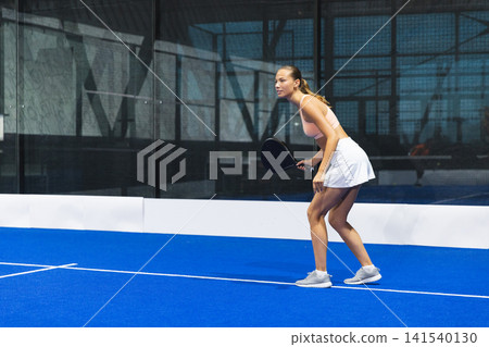 Asian female athlete crouching on blue padel court at gym holding black padel racket, copy space Asian female athlete crouching on blue padel court at gym holding black padel racket, copy space 141540130