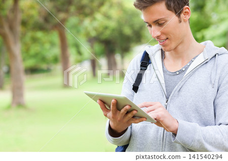 Man in twenties standing right in park using tablet, in gray hoodie and backpack, copy space Man in twenties standing right in park using tablet, in gray hoodie and backpack, copy space 141540294