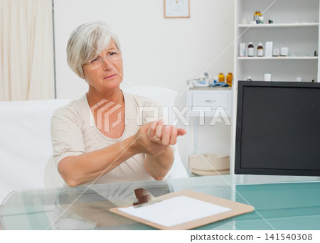 Senior female holding wrist while consulting in medical exam room with clipboard on glass desk 141540308