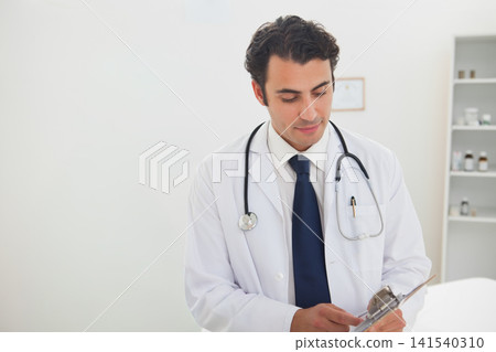 Mid-adult male physician in clinic in lab coat holding clipboard and reviewing chart, copy space Mid-adult male physician in clinic in lab coat holding clipboard and reviewing chart, copy space 141540310