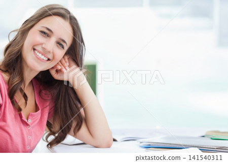 Adult female sitting at desk in pink top smiling with notebooks papers books, copy space Adult female sitting at desk in pink top smiling with notebooks papers books, copy space 141540311