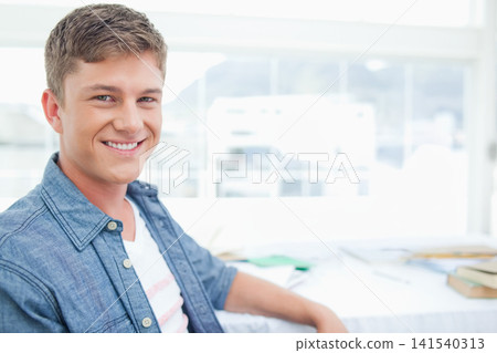 Man in twenties sitting at desk in bright study wearing denim shirt, papers, books, copy space Man in twenties sitting at desk in bright study wearing denim shirt, papers, books, copy space 141540313