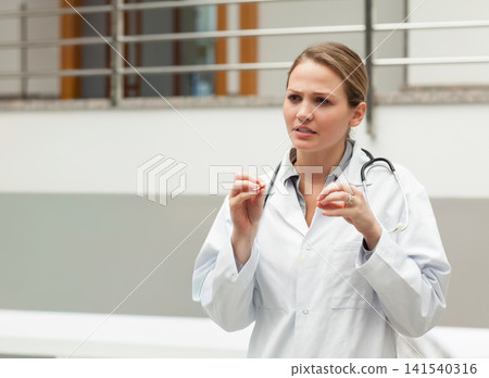 Female doctor standing in clinic hallway pinching gesture in lab coat with stethoscope, copy space Female doctor standing in clinic hallway pinching gesture in lab coat with stethoscope, copy space 141540316