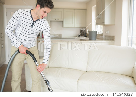 Man vacuuming cream leather sofa with canister vacuum wand and hose at home, copy space 141540328