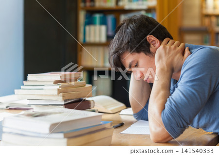 Male student in blue shirt sitting at library table leaning over notebook, books nearby, copy space Male student in blue shirt sitting at library table leaning over notebook, books nearby, copy space 141540333