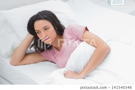Female reclining on bed in bedroom, holding white duvet, resting on pillows, wearing pink T-shirt Female reclining on bed in bedroom, holding white duvet, resting on pillows, wearing pink T-shirt 141540346