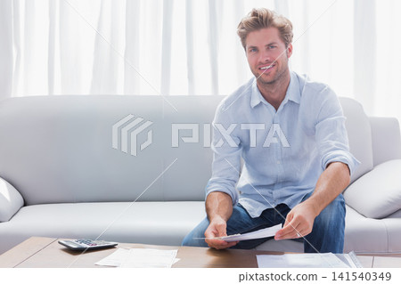 Mid-adult male wearing blue shirt, sitting right on sofa sorting papers with calculator, copy space Mid-adult male wearing blue shirt, sitting right on sofa sorting papers with calculator, copy space 141540349
