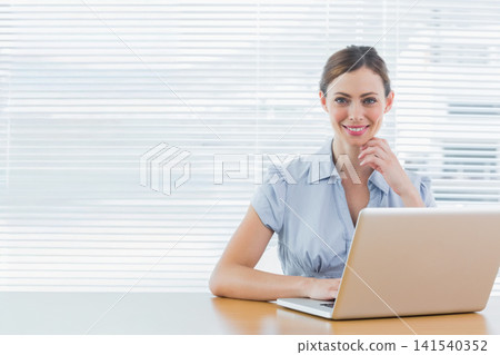 Mid-adult woman working at silver laptop wearing light blue blouse with venetian blinds, copy space Mid-adult woman working at silver laptop wearing light blue blouse with venetian blinds, copy space 141540352