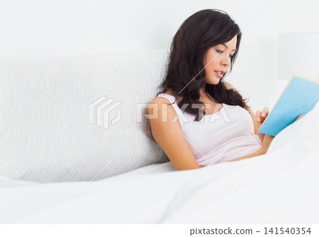 Woman reclining propped against quilted headboard on bed, reading blue paperback book, copy space Woman reclining propped against quilted headboard on bed, reading blue paperback book, copy space 141540354