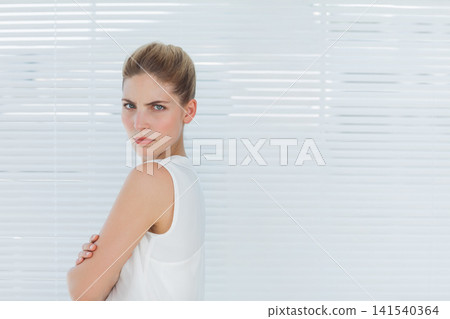 Mid 20s woman standing arms crossed, wearing white tank top before horizontal blinds, copy space 141540364