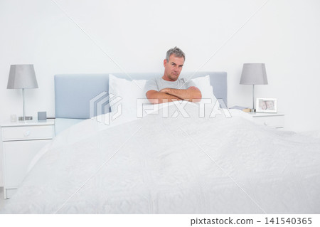 Senior man sitting against blue headboard on white bed between nightstands, wearing grey T-shirt Senior man sitting against blue headboard on white bed between nightstands, wearing grey T-shirt 141540365