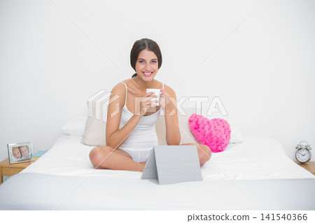Female sitting cross-legged on bed in white tank top and shorts, holding mug, tablet visible Female sitting cross-legged on bed in white tank top and shorts, holding mug, tablet visible 141540366