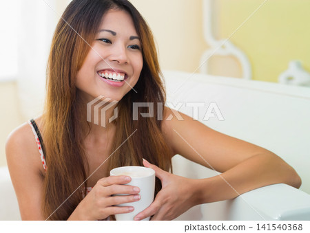 Adult Asian woman sitting on white couch at home holding mug and smiling in sleeveless top Adult Asian woman sitting on white couch at home holding mug and smiling in sleeveless top 141540368