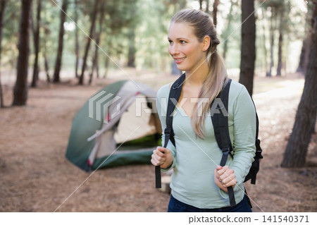 Female hiker wearing light green top standing in pine campsite holding backpack near green tent Female hiker wearing light green top standing in pine campsite holding backpack near green tent 141540371