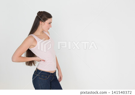 Female model posing profile left white backdrop wearing pink tank, jeans, stud earrings, copy space 141540372